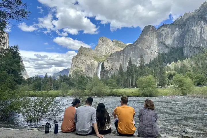 Five people sitting by a river facing mountains and waterfall.