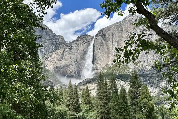 Waterfall flowing down a cliff surrounded by trees under a blue sky with clouds.